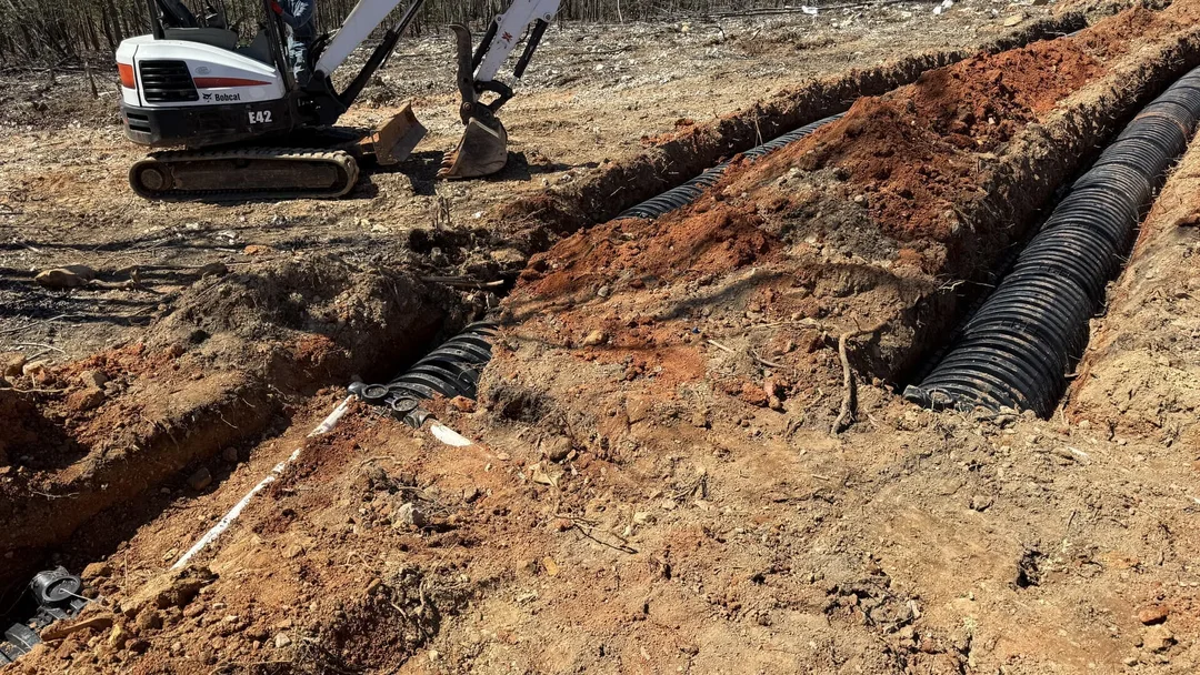 Bobcat E42 mini excavator digging trenches for two black corrugated pipes in red dirt.
